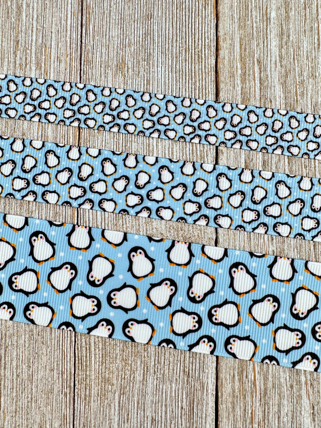 May include: Three ribbons with a penguin pattern against a light blue background. The top ribbon has small penguins, the middle has medium-sized penguins, and the bottom ribbon has larger penguins. The penguins are black and white with orange feet and beaks. The ribbons are on a wooden surface.