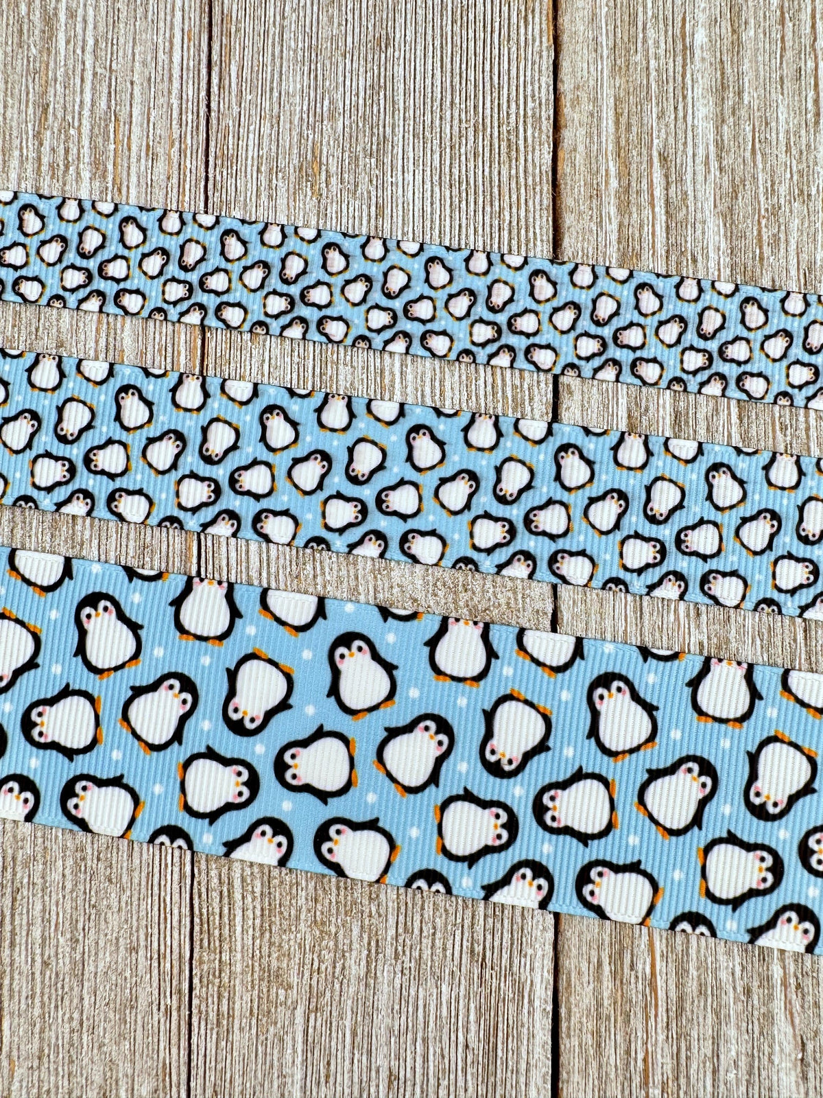 May include: Three ribbons with a penguin pattern against a light blue background. The top ribbon has small penguins, the middle has medium-sized penguins, and the bottom ribbon has larger penguins. The penguins are black and white with orange feet and beaks. The ribbons are on a wooden surface.