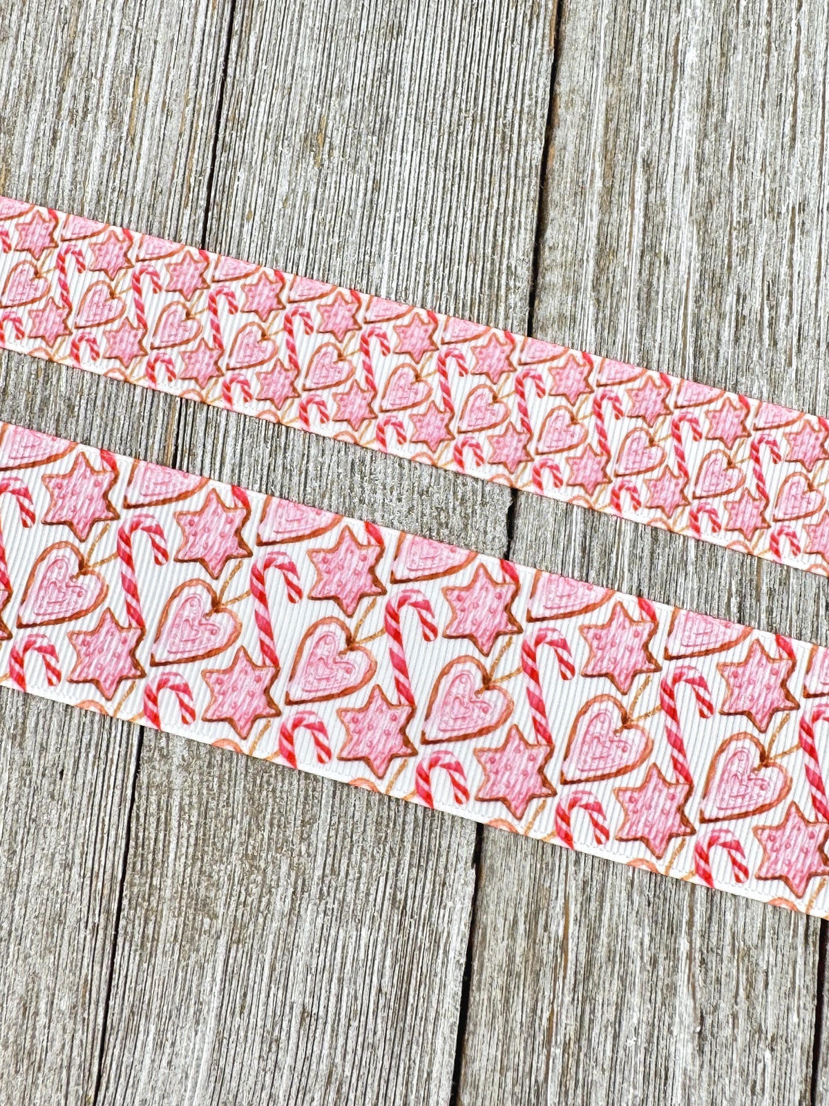 May include: Two ribbons with a repeating pattern of pink gingerbread cookies, candy canes, and star-shaped cookies on a white background. The cookies have a brown outline and the candy canes have red and white stripes. The ribbons are displayed on a wooden surface.