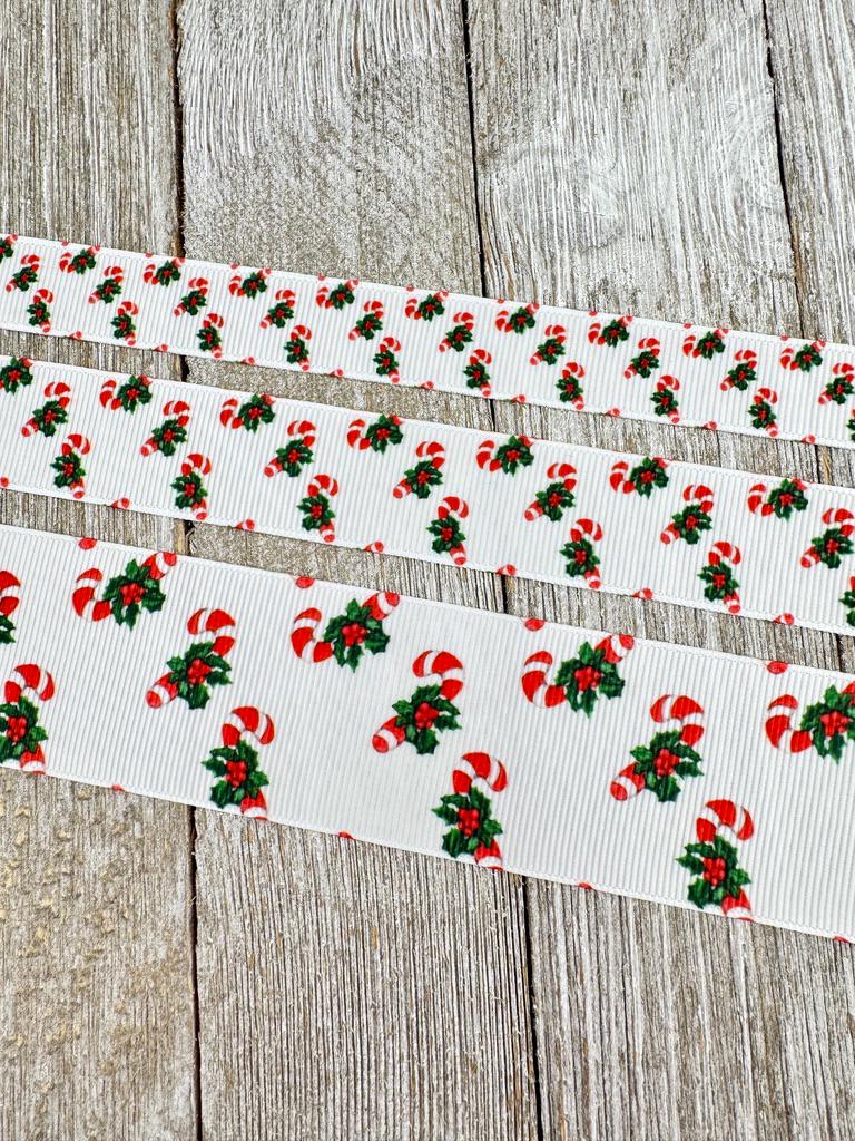 May include: Three white ribbons with a repeating pattern of red and white candy canes and green holly leaves. The ribbons vary in width, with the widest ribbon at the bottom. The candy canes have red stripes and are paired with green holly and red berries, creating a festive Christmas theme.