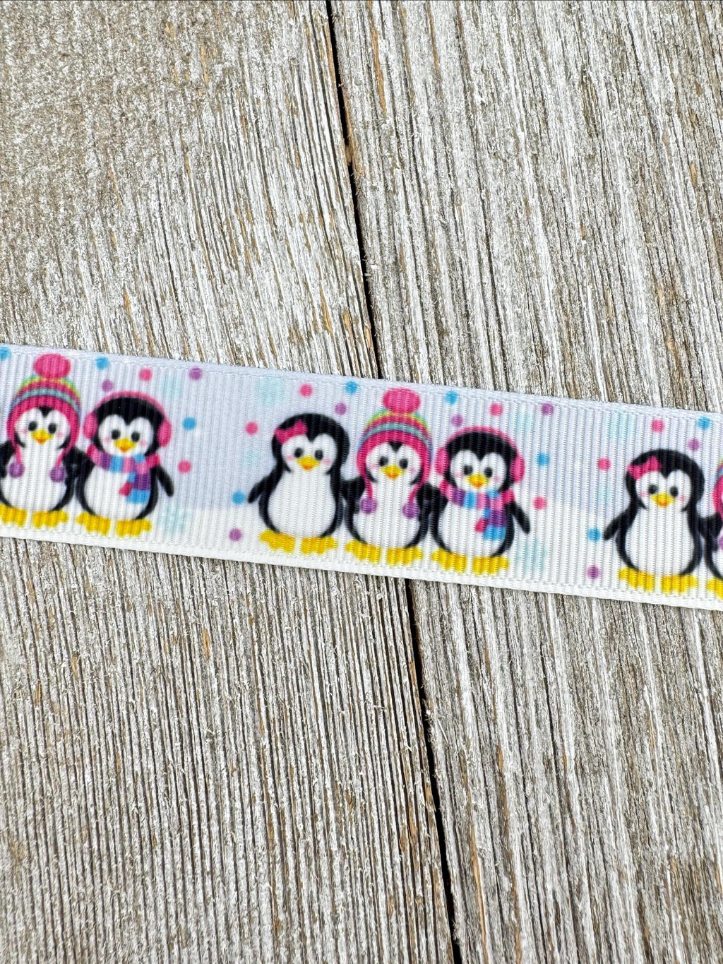 May include: A white ribbon with a repeating pattern of cartoon penguins. The penguins are black and white with yellow feet and beaks, wearing hats and scarves in pink, blue, and purple. Small pink and purple polka dots are scattered between the penguins.