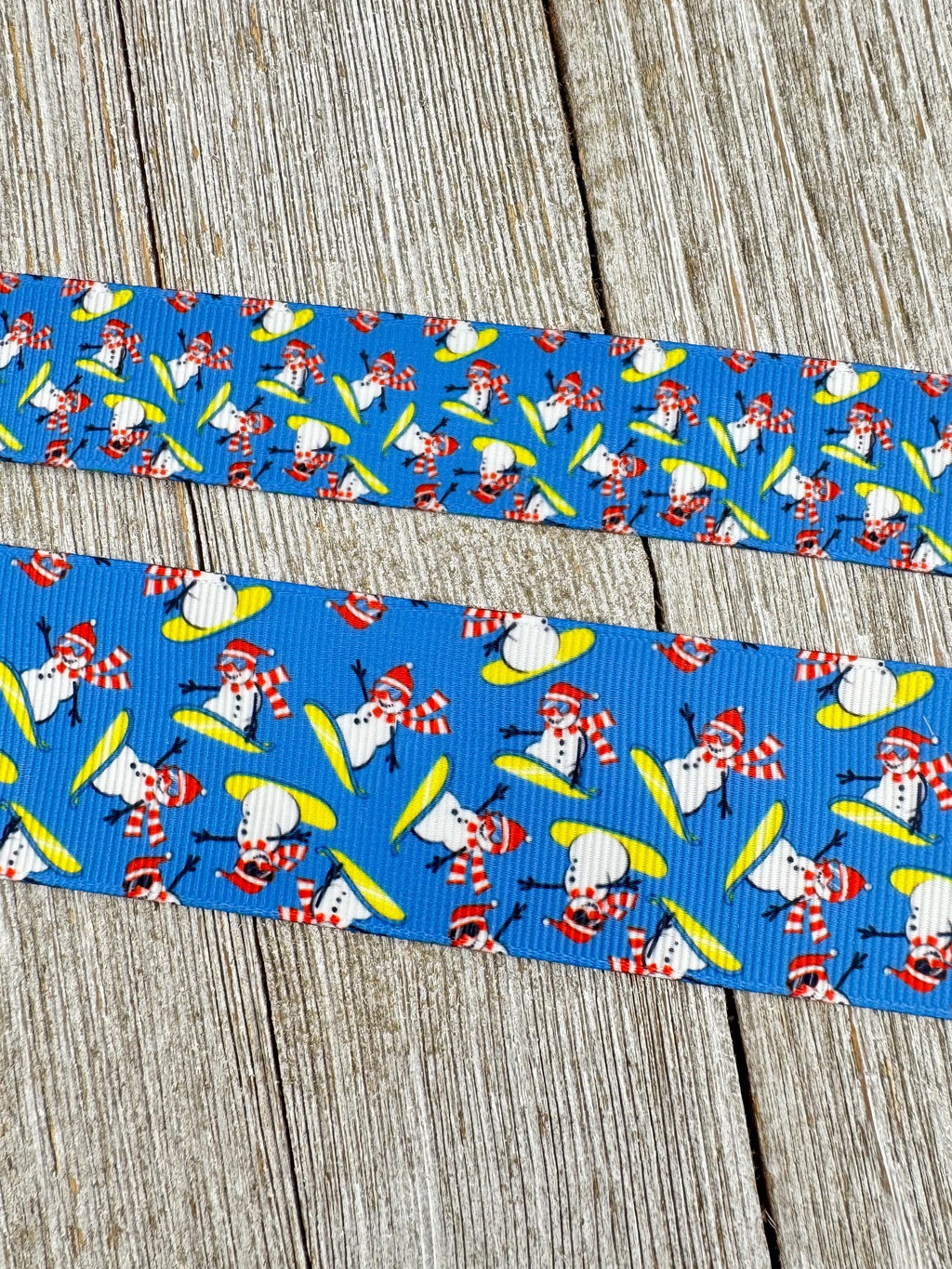 May include: Two strips of blue ribbon with a repeating pattern of snowmen on skis. The snowmen are white with red hats, scarves, and sunglasses. They are skiing on yellow skis. The ribbon is on a wooden surface.