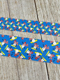 May include: Two strips of blue ribbon with a repeating pattern of snowmen on skis. The snowmen are white with red hats, scarves, and sunglasses. They are skiing on yellow skis. The ribbon is on a wooden surface.