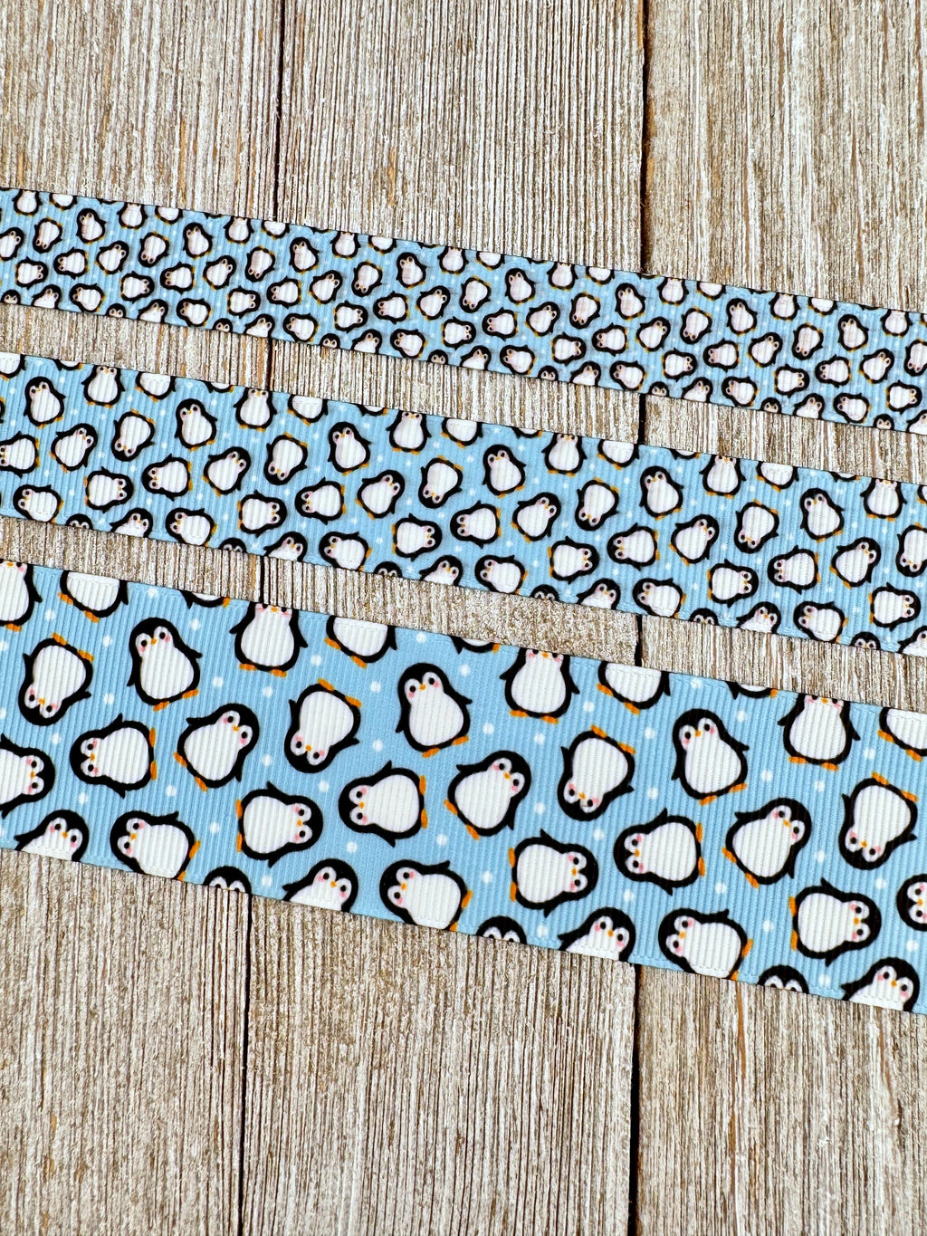 May include: Three ribbons with a penguin pattern on a light blue background. The top ribbon has small penguins, the middle ribbon has medium-sized penguins, and the bottom ribbon has large penguins. The penguins are white with black details and orange beaks.