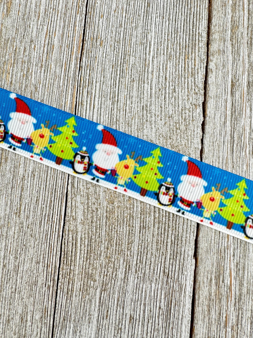 May include: A blue ribbon featuring a repeating Christmas pattern. The pattern includes Santa Claus in a red hat and white beard, reindeer with yellow antlers, green Christmas trees, and penguins. The ribbon is set against a wooden background.