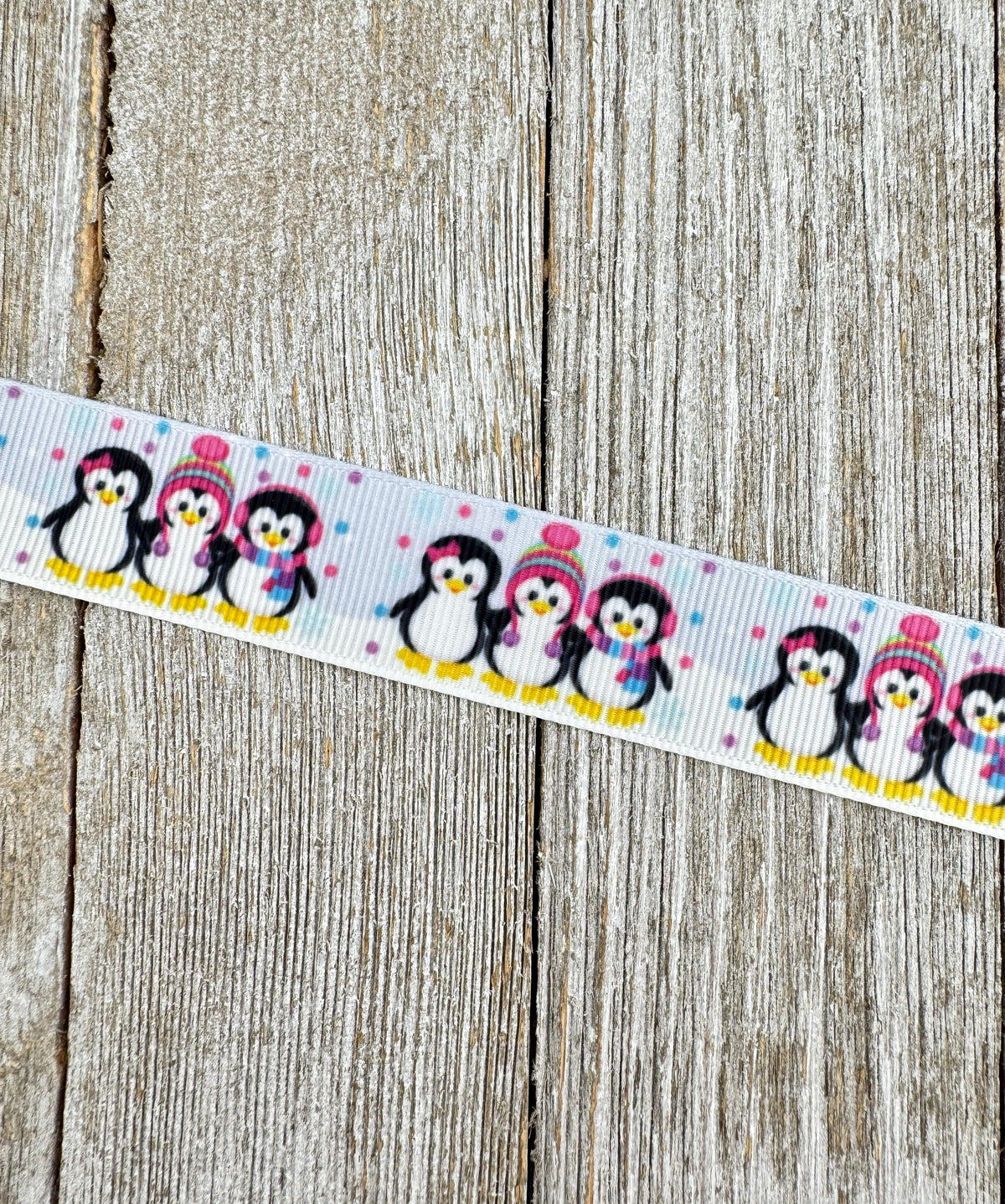 May include: A white ribbon with a repeating pattern of penguins. Each penguin is black and white with a yellow beak and feet. Some penguins wear pink and red hats and scarves. The ribbon also features small pink and blue polka dots.