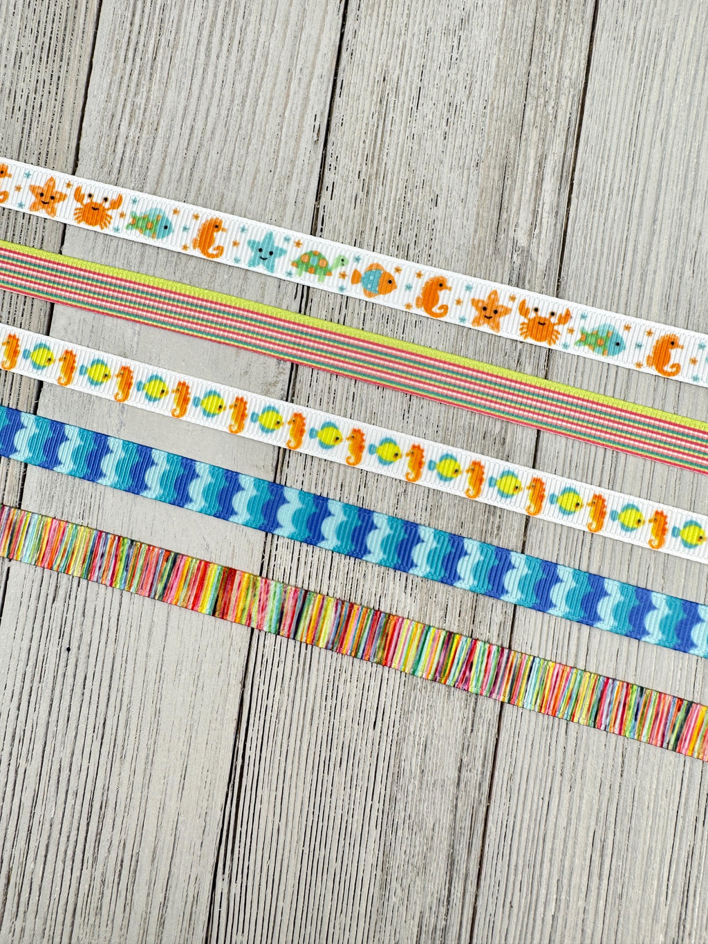 May include: Five strips of decorative ribbon arranged horizontally. The top ribbon features colorful sea creatures on a white background. The second has thin multicolored stripes. The third displays yellow fish and orange seahorses. The fourth has blue waves. The bottom ribbon features vertical multicolored stripes.