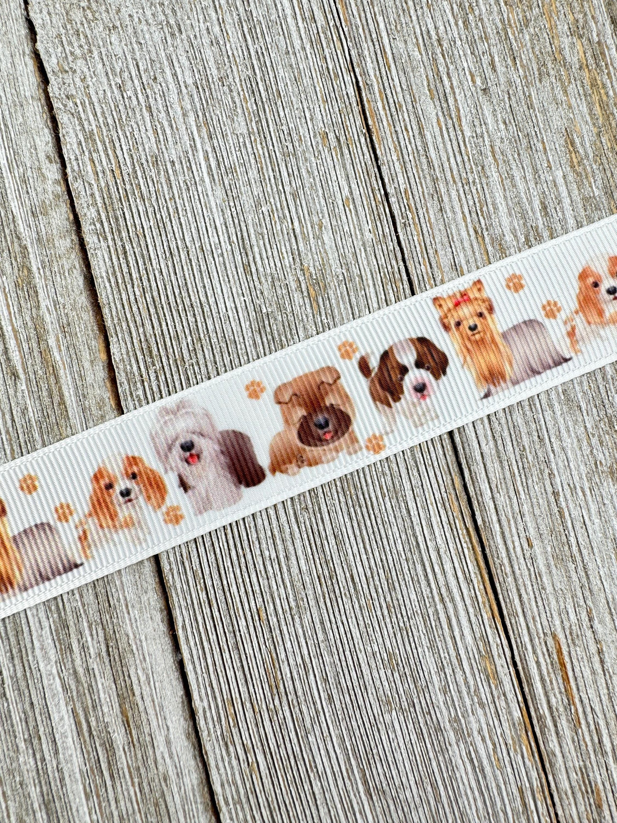 May include: White ribbon featuring a repeating pattern of various dog breeds in shades of brown, tan, and white. Small paw prints are interspersed between the dog illustrations. The ribbon is set against a weathered, light-colored wooden surface.